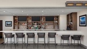 A bar area with a white countertop, multiple shelves of bottles, and six wicker bar stools. - Hilton Garden Inn Santa Barbara / Goleta (Goleta)