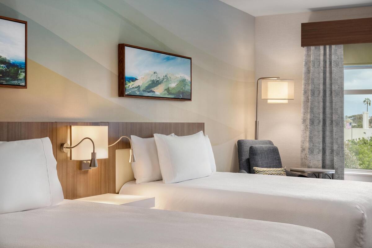 Une chambre d’hôtel avec deux lits, un bureau, une chaise, une lampe et une fenêtre avec des rideaux.