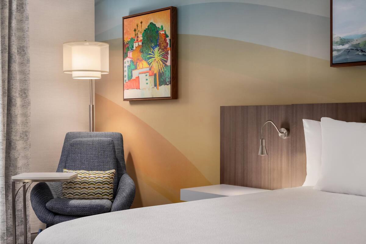 Une chambre d’hôtel avec un lit, une chaise, une table, une lampe et un tableau au mur.