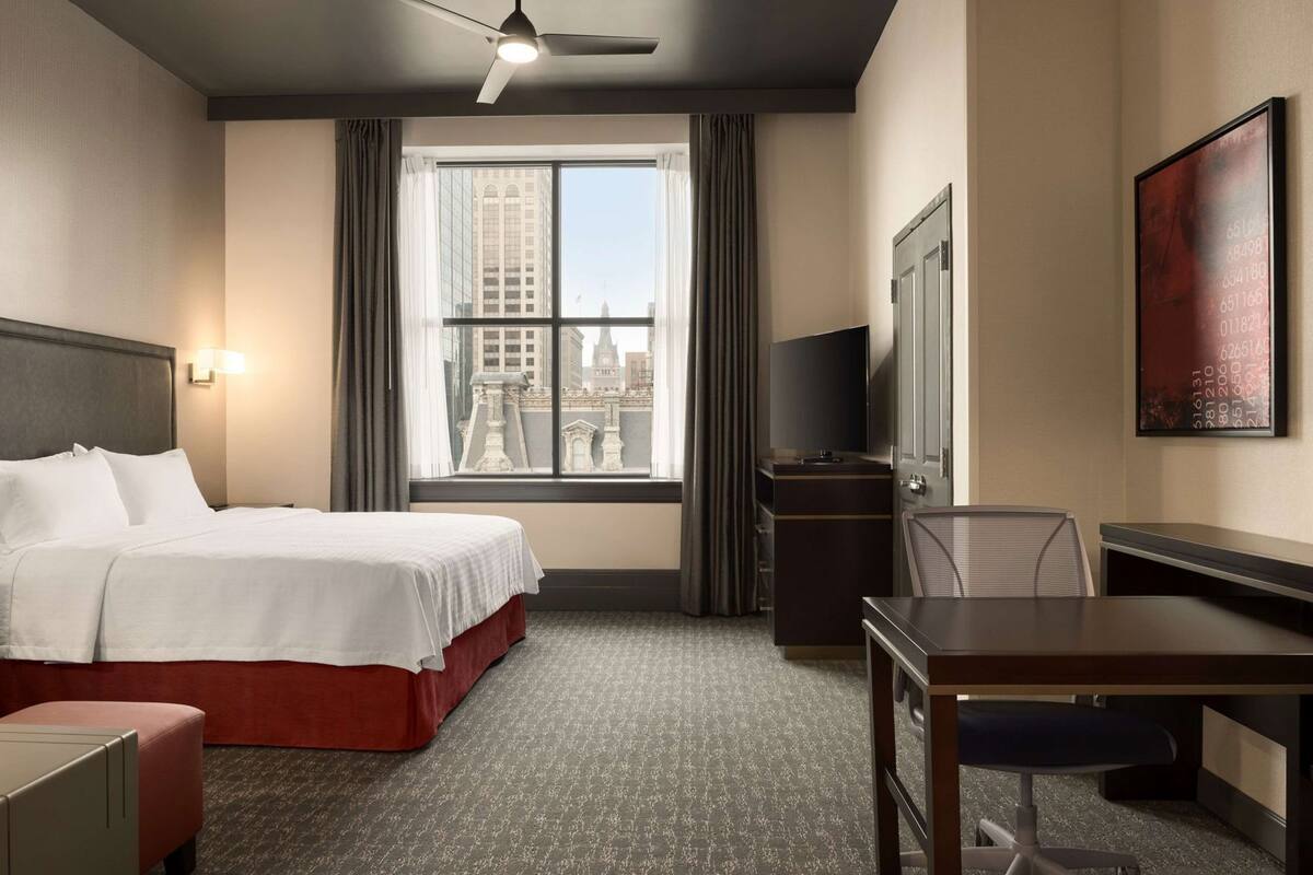Ein Hotelzimmer mit Bett, Schreibtisch, Stuhl, Fernseher und einem Fenster mit Blick auf die Stadt.