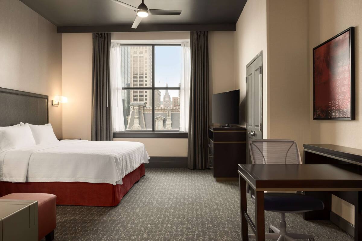 Ein Hotelzimmer mit einem Bett, einem Schreibtisch, einem Stuhl, einem Fernseher und einem Fenster mit Blick auf die Stadt.