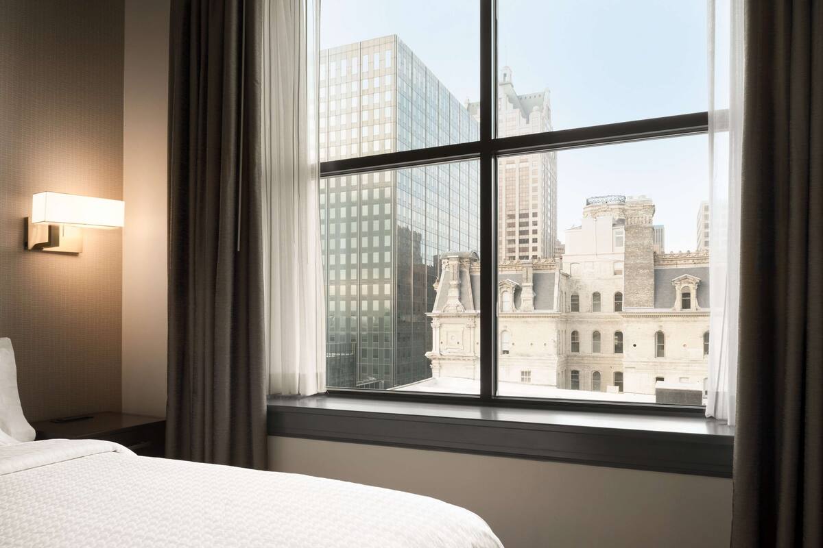 Ein Hotelzimmer mit großem Fenster, einem Bett, einem Nachttisch und einer Wandleuchte.