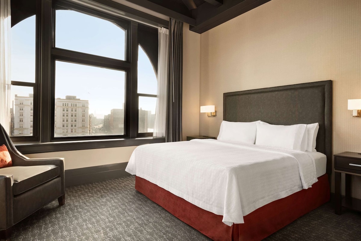 A hotel room with a large bed, a chair, a nightstand, and a window with a city view.
