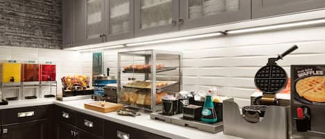 A breakfast bar with a waffle maker, a coffee machine, and a variety of food items.
