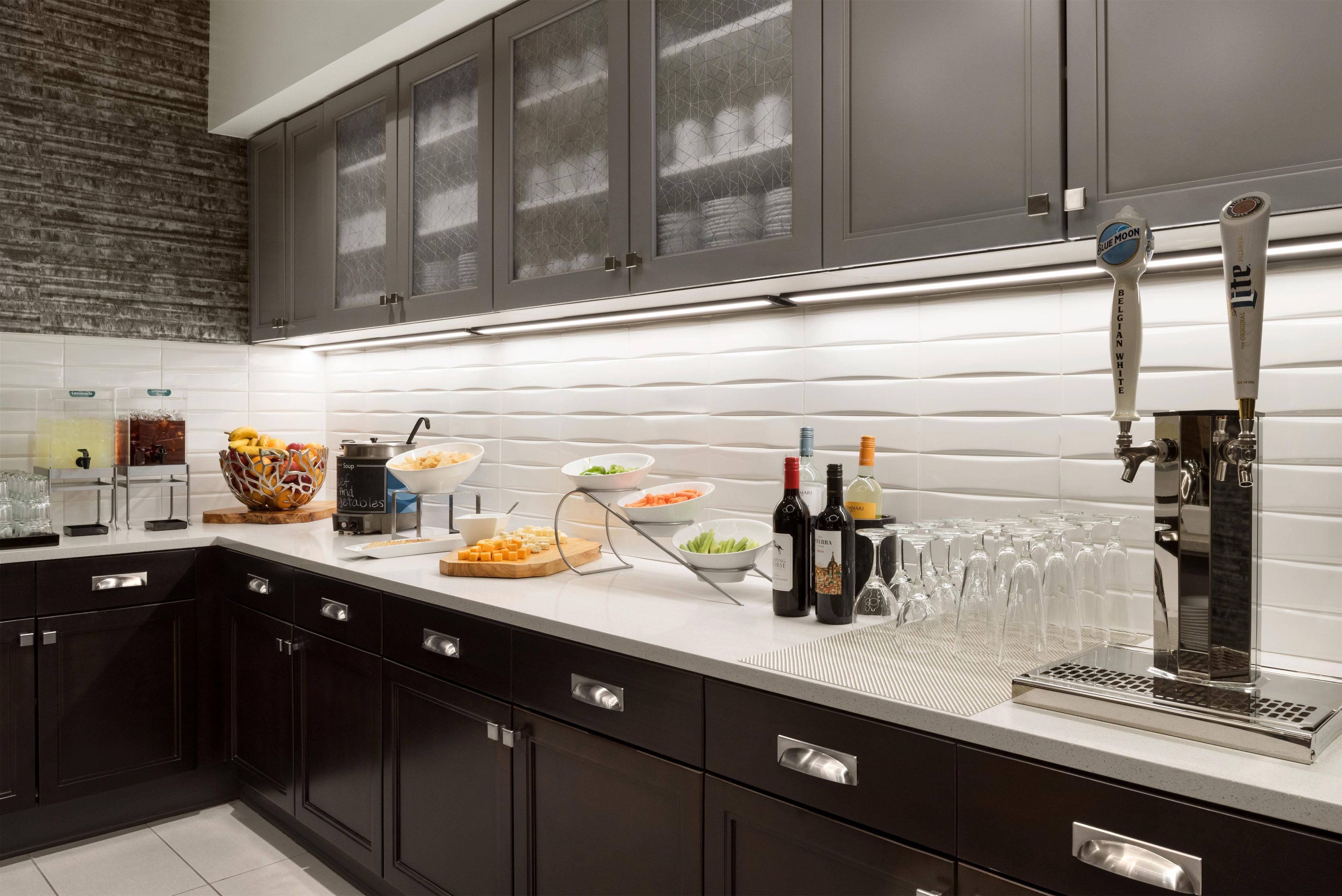 Una cocina moderna con armarios oscuros, encimera blanca y una variedad de alimentos y bebidas.