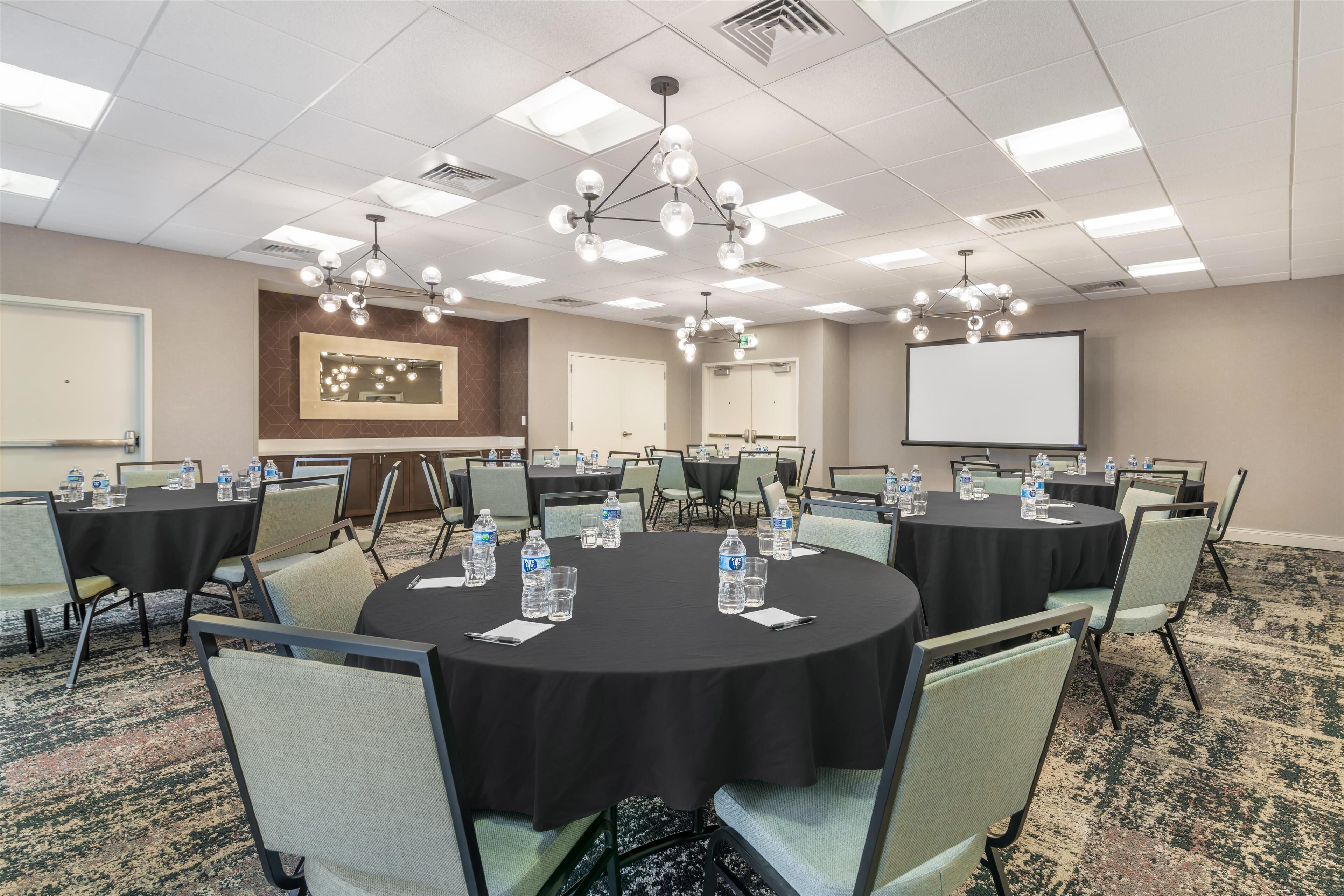 A conference room with round tables, chairs, water bottles, a projector screen, and a large mirror.