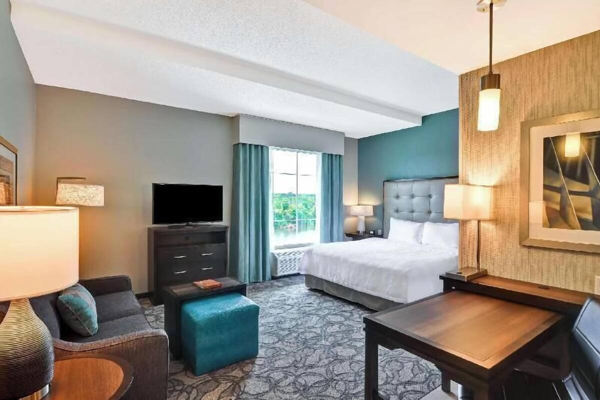 A hotel room with a bed, a TV, a desk, a chair, a sofa, and a window with curtains.