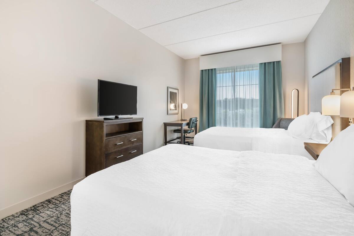 A hotel room with two beds, a desk with a chair, a flat-screen TV, a lamp, and a window with curtains.