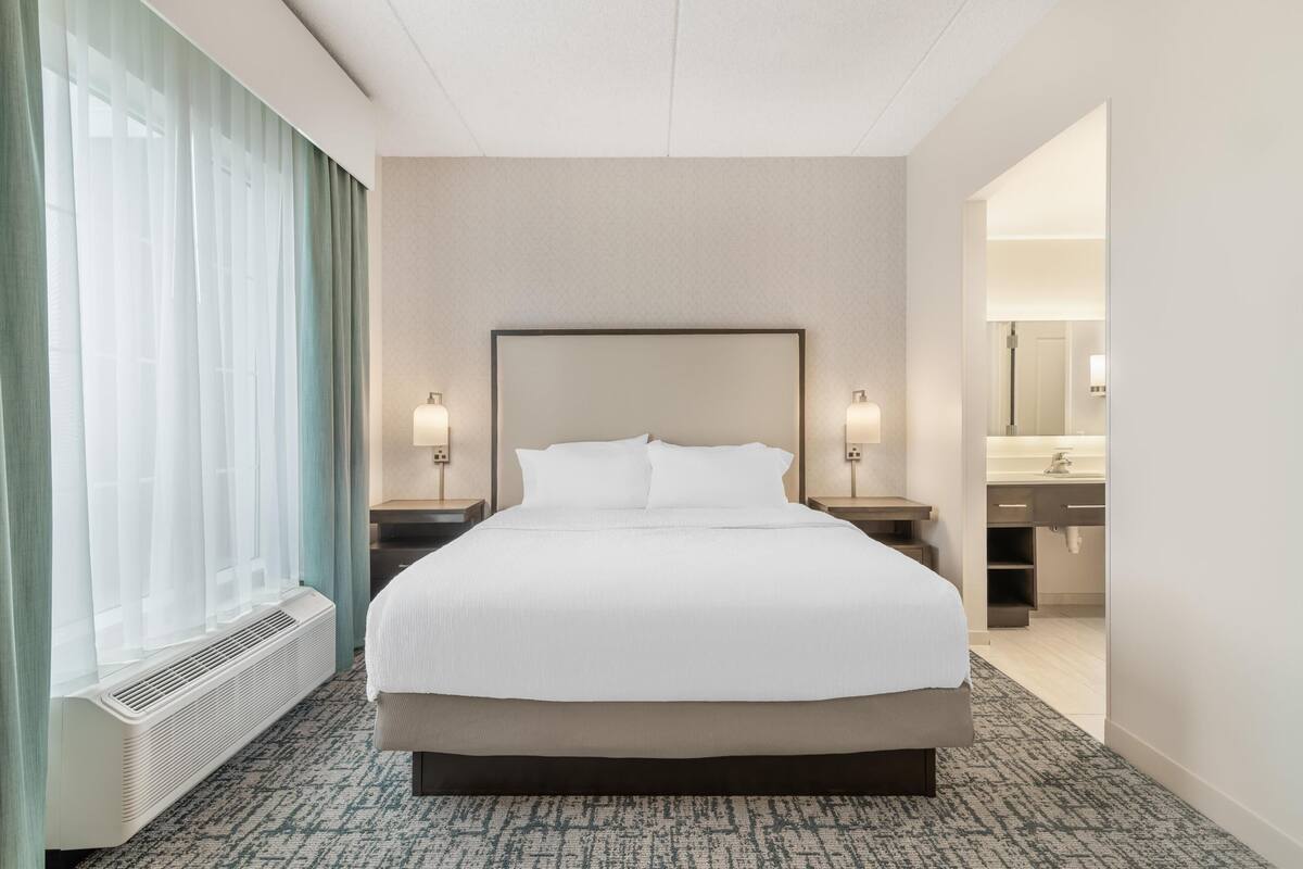 A hotel room with a large bed, two bedside tables with lamps, a window with curtains, and a view into the bathroom.