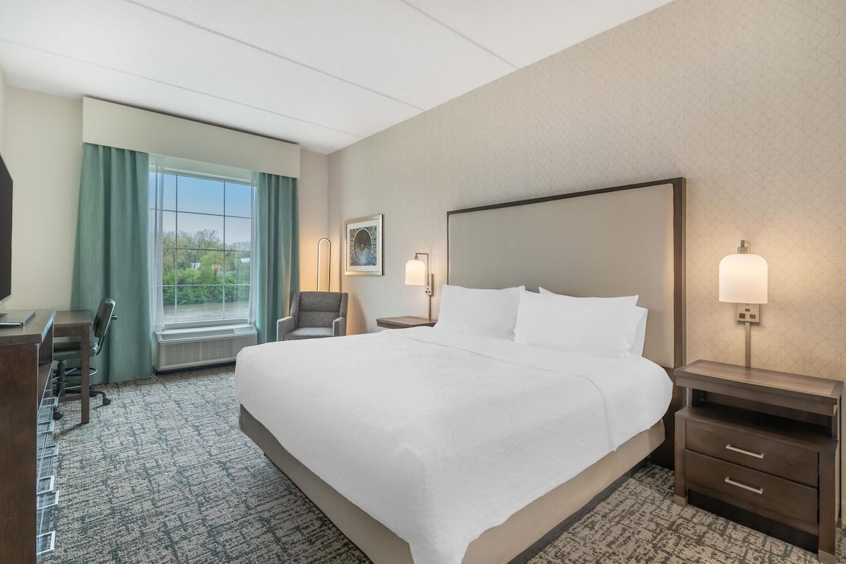 A hotel room with a large bed, a desk, and a chair. There is a window with curtains, a picture on the wall, and a nightstand with a lamp.
