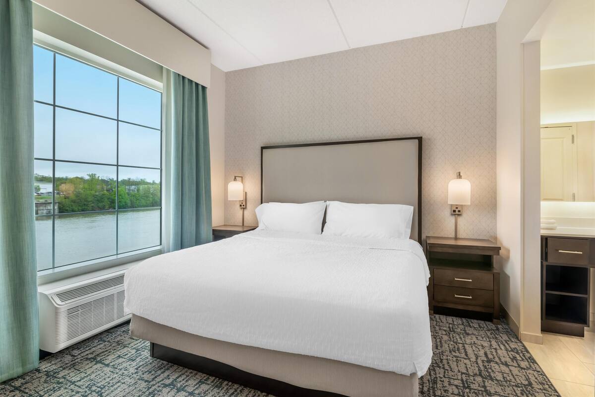 A hotel room with a large bed, two bedside tables with lamps, a window with a view of greenery, and a bathroom with a mirror and sink.