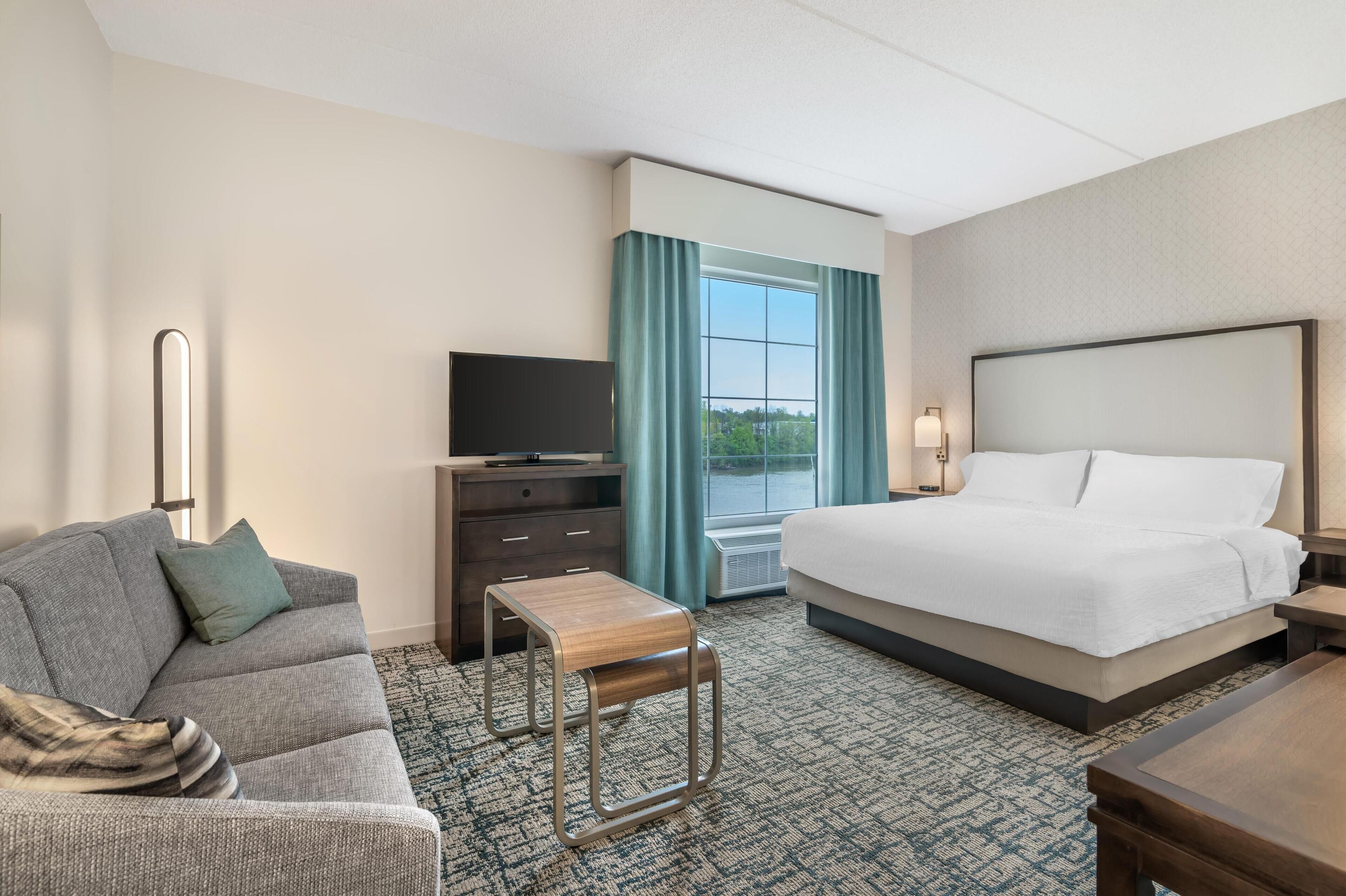 A hotel room with a bed, a sofa, a small wooden stool, a TV, and a desk.