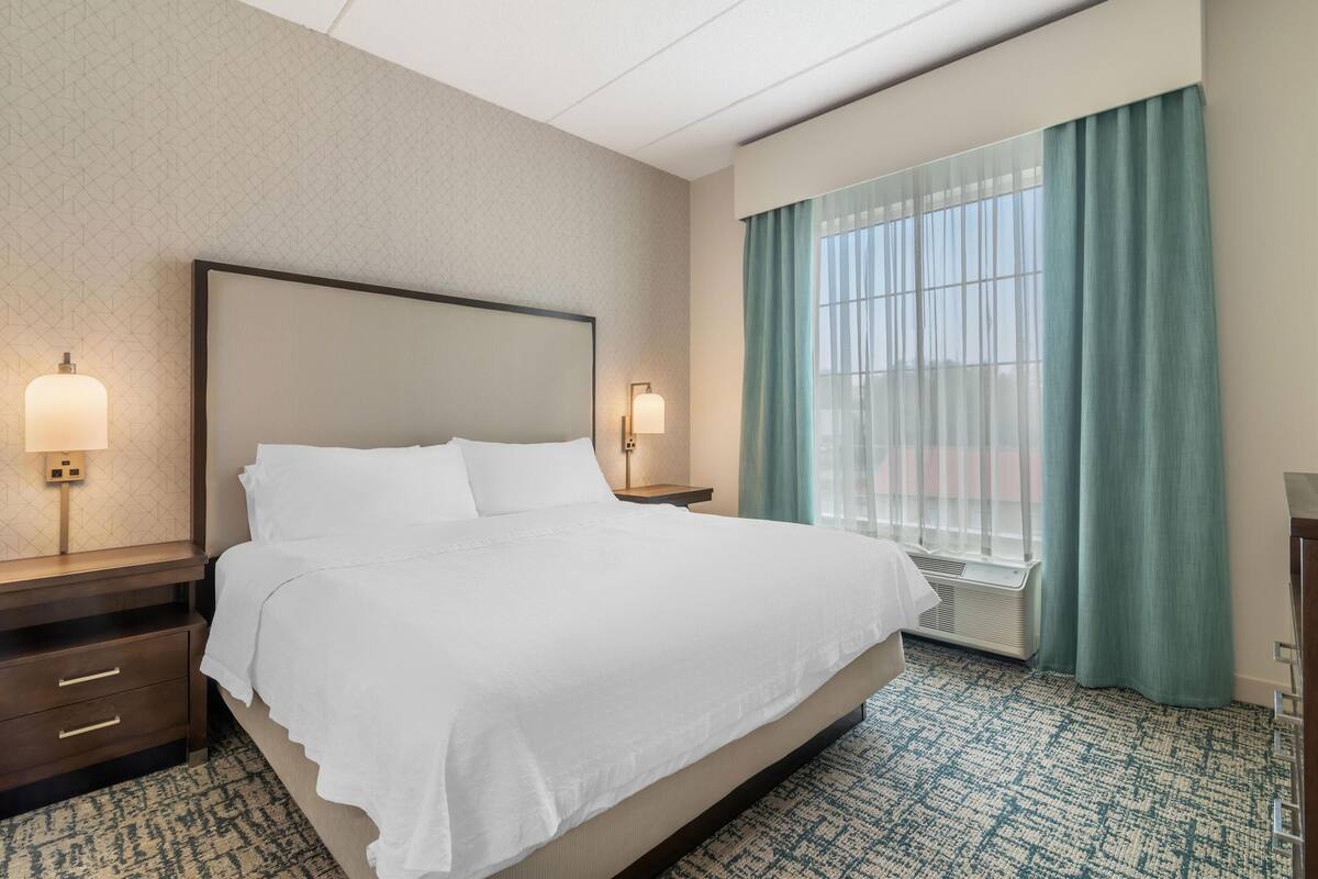 A hotel room with a large bed, two bedside tables with lamps, a window with curtains, and a patterned carpet.