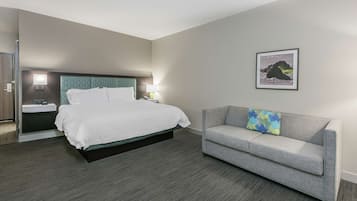 A hotel room with a bed, a gray sofa, a framed picture on the wall, and a nightstand with a lamp.
