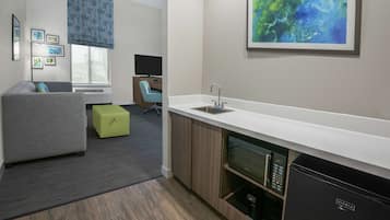 A modern office space with a kitchenette, a grey sofa, a green ottoman, and a large abstract painting on the wall.