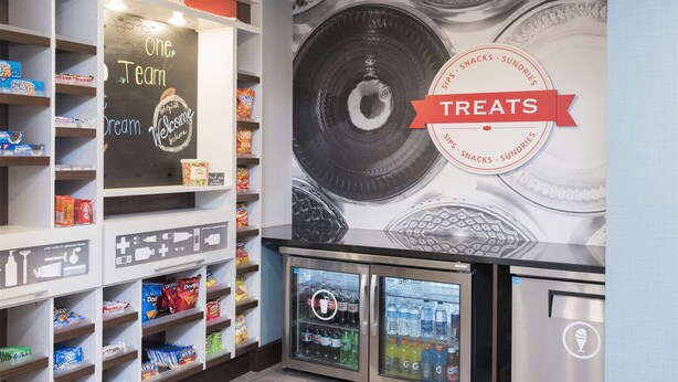 A snack and sundries display with a chalkboard sign reading "One Team Dream Welcome" and a refrigerator stocked with beverages. - Hampton Inn Westfield (Westfield)