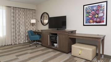 Une chambre d’hôtel avec un bureau en bois, une télévision, une chaise bleue, une valise, une lampe et un tableau au mur.