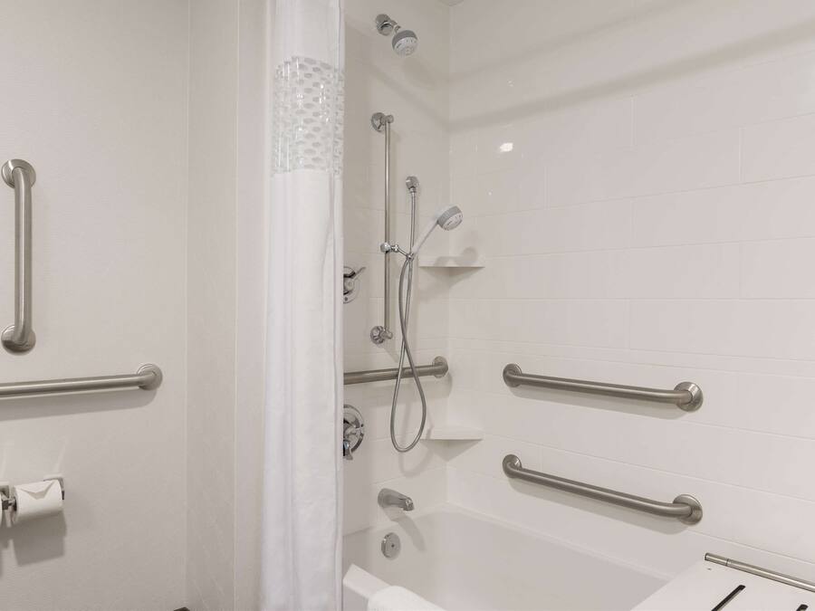 Baño con azulejos blancos, ducha con cabezal de ducha manual y barras de apoyo.