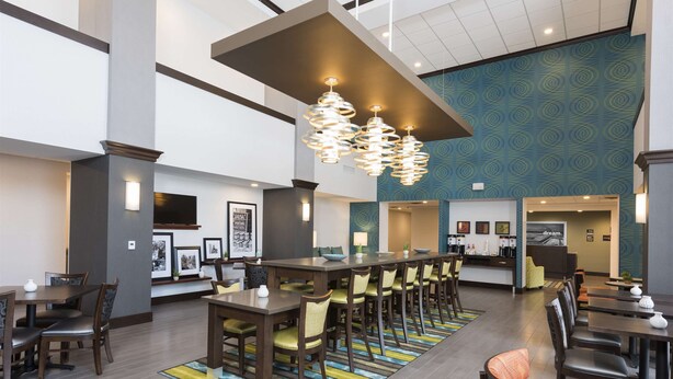 A hotel lobby with a long reception desk, multiple dining tables with chairs, and a patterned wall. - Hampton Inn Westfield (Westfield)