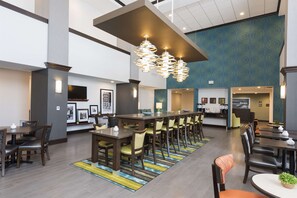 A hotel lobby with a long reception desk, multiple dining tables with chairs, and a patterned wall.