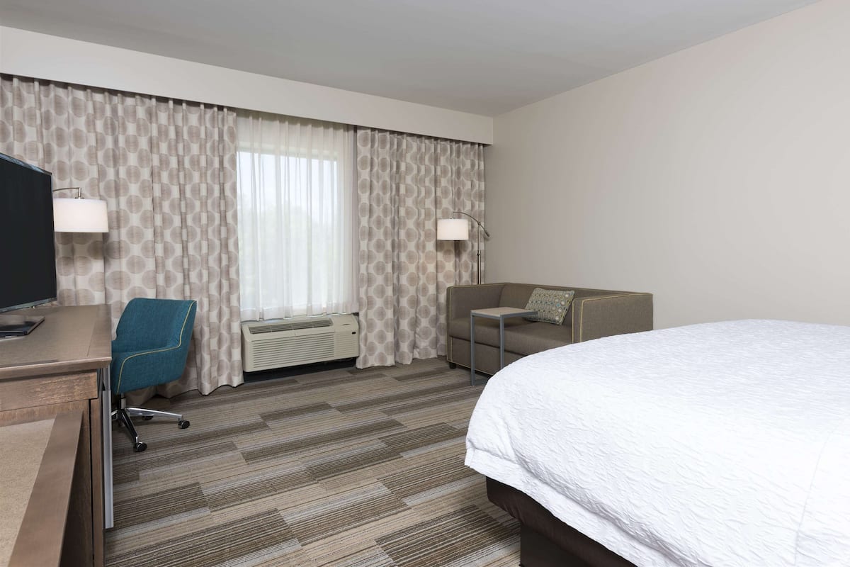 Quarto de hotel com cama, escrivaninha com televisão, cadeira de escritório azul, sofá e duas luminárias.