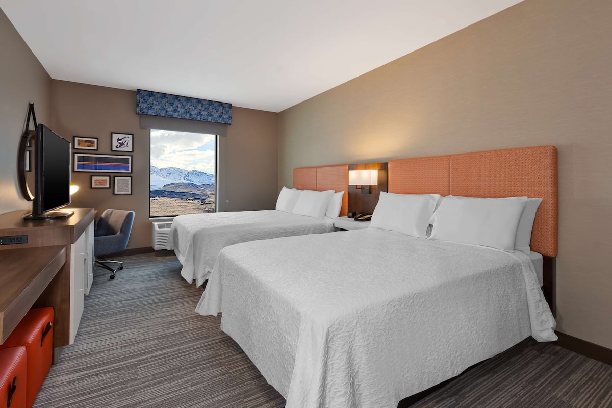 A hotel room with two beds, a desk, a chair, a TV, and a window with a mountain view.