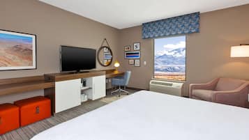 A hotel room with a large bed, a desk with a computer, a TV, and a view of mountains through the window.