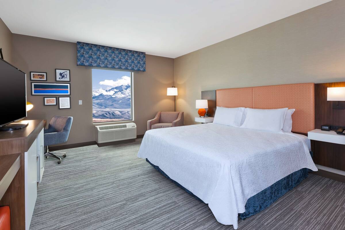 A hotel room with a large bed, a desk with a computer, a chair, a window with a mountain view, and a wall with framed pictures.