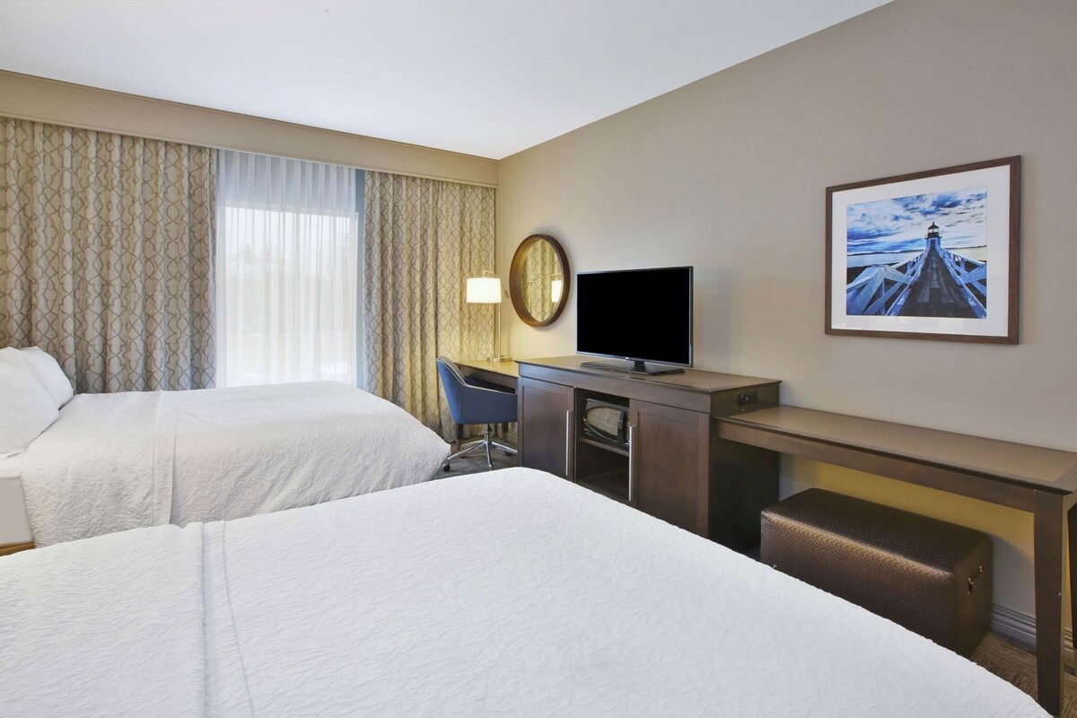 Habitación de hotel con dos camas, un escritorio con televisor, una silla, lámpara de escritorio, reloj y un cuadro en la pared.