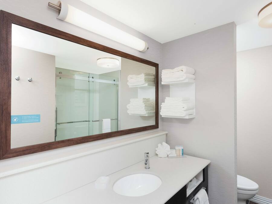 A bathroom with a large mirror, a sink, and a toilet. There are towels stacked on a shelf.