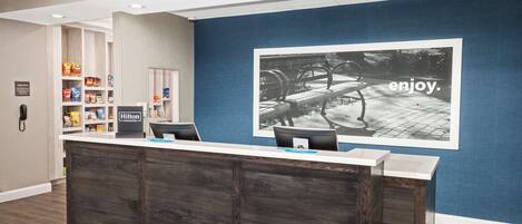 A reception area with a wooden reception desk, a blue wall with a large photo, and shelves stocked with snacks and toiletries.