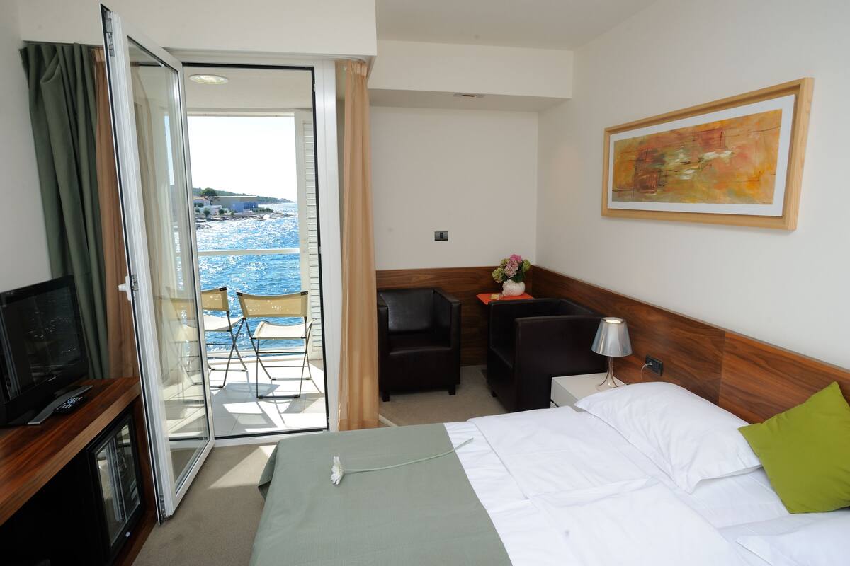 A hotel room with a bed, a desk, a chair, a TV, and a view of the sea.