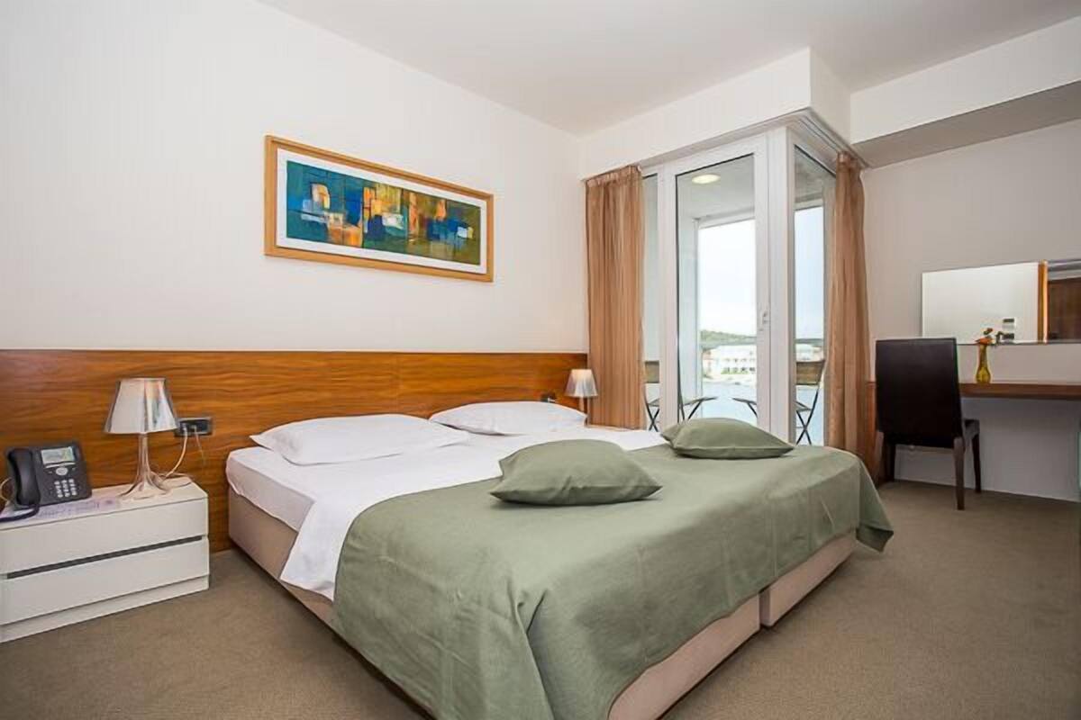 A hotel room with a bed, bedside table with a phone, a desk with a chair, a painting on the wall, and a view through a glass door.