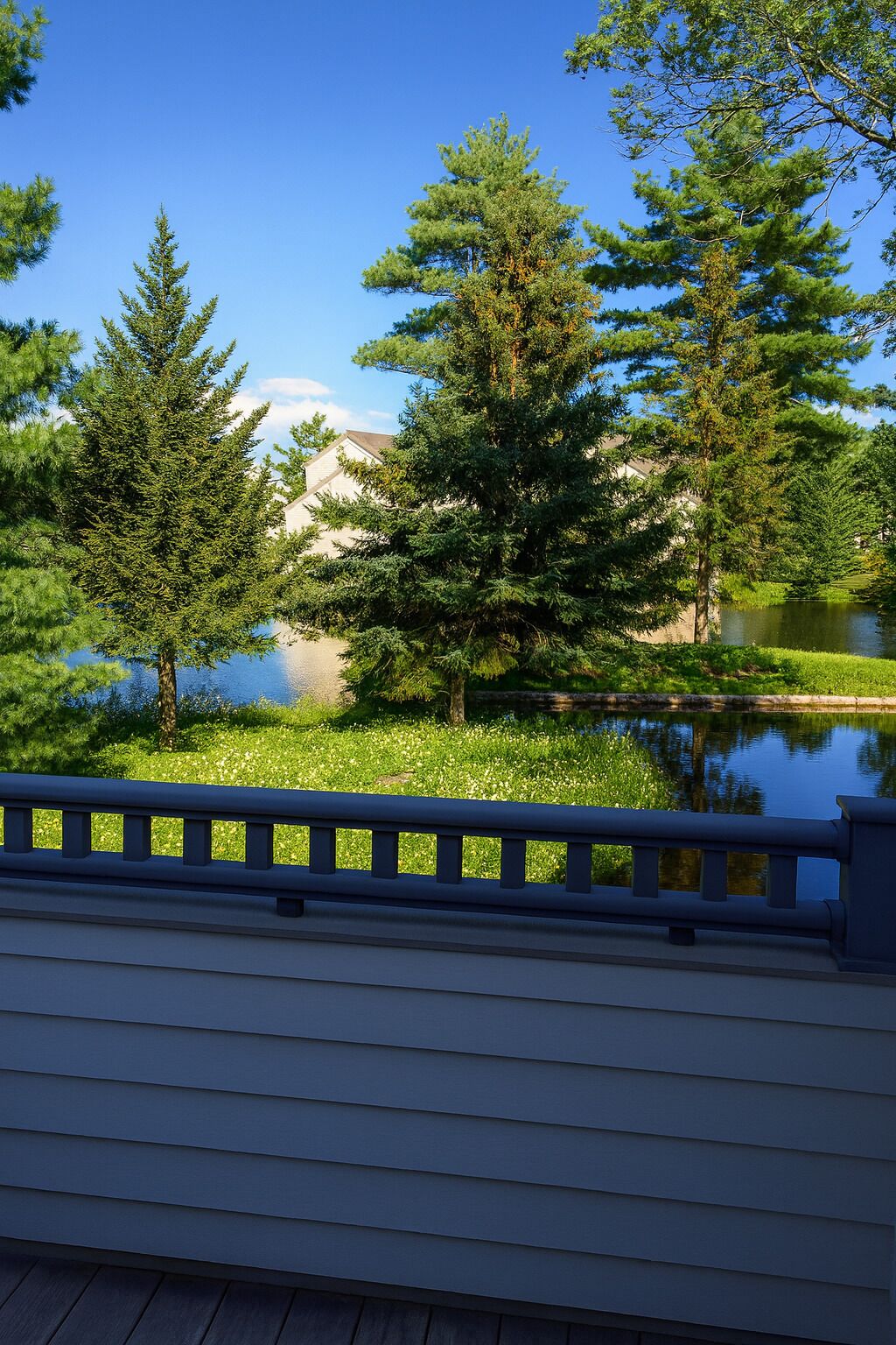 A wooden deck with a railing overlooking a calm lake, surrounded by lush greenery and tall trees.
