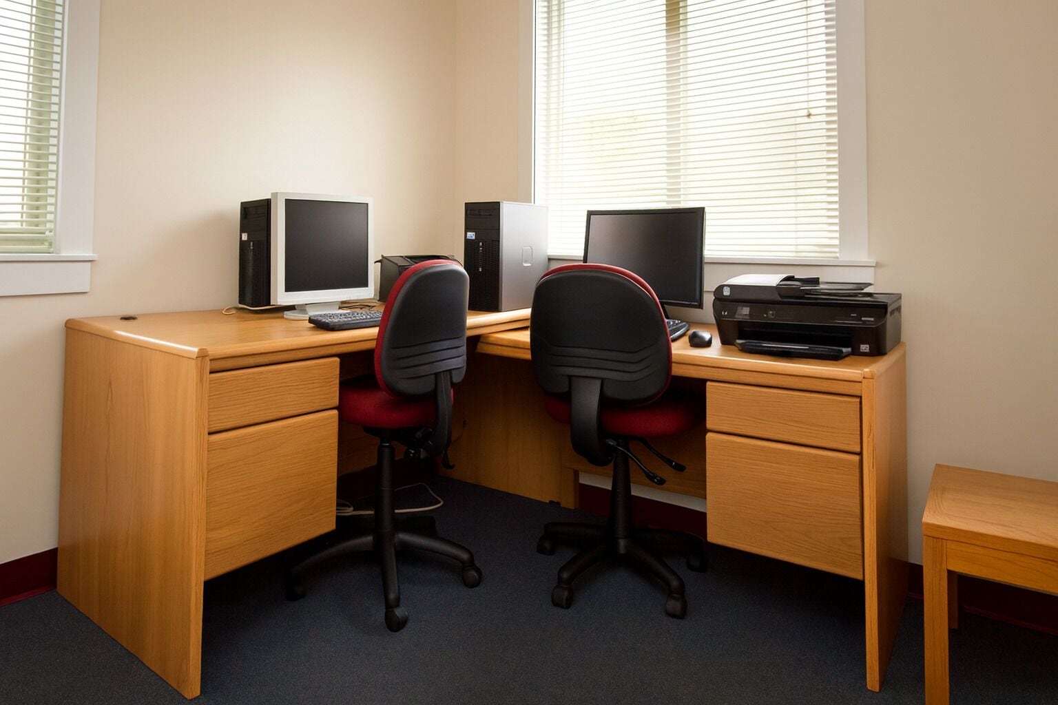 A corner office with two desks, each with a computer monitor, keyboard, and printer. There are two office chairs and a small wooden table.