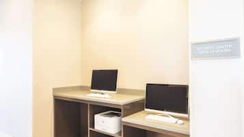 Un centro de negocios con dos estaciones de trabajo, cada una con monitor, teclado y ratón. Hay una impresora en el suelo. Un cartel indica que el centro de negocios está abierto las 24 horas.