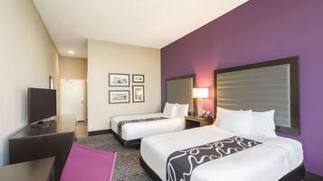 A hotel room with two beds, a desk, a TV, and a door leading to a hallway with framed pictures.
