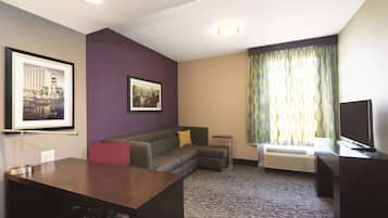 A hotel room with a desk, a sofa, a framed cityscape picture, a window with curtains, and a flat-screen TV.
