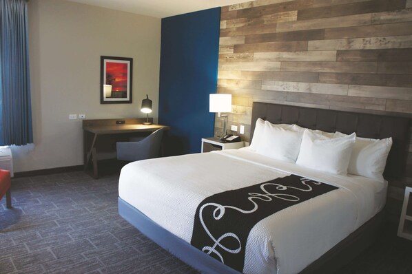 A hotel room with a large bed, a desk with a lamp, a chair, and a wall with a wooden panel design. - La Quinta Inn & Suites by Wyndham Pittsburg (Pittsburg)