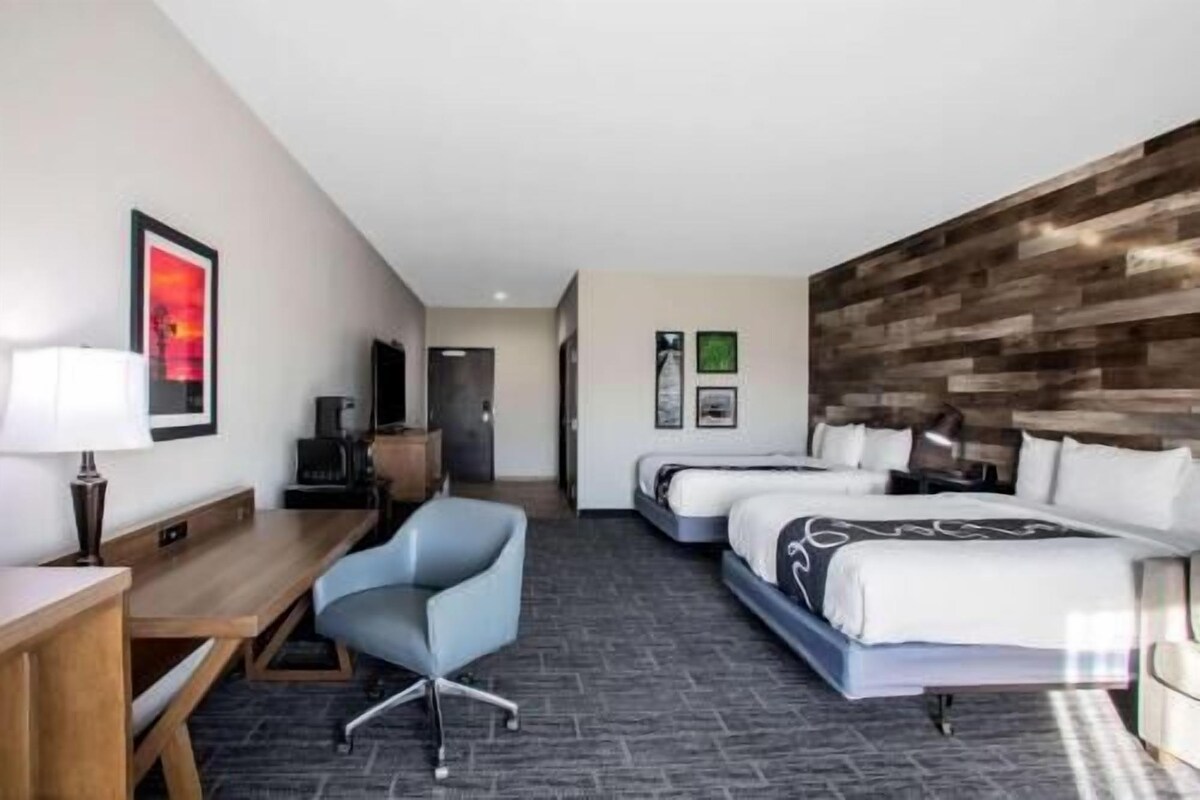 A hotel room with a desk, two beds, a chair, a TV, and artwork on the walls.