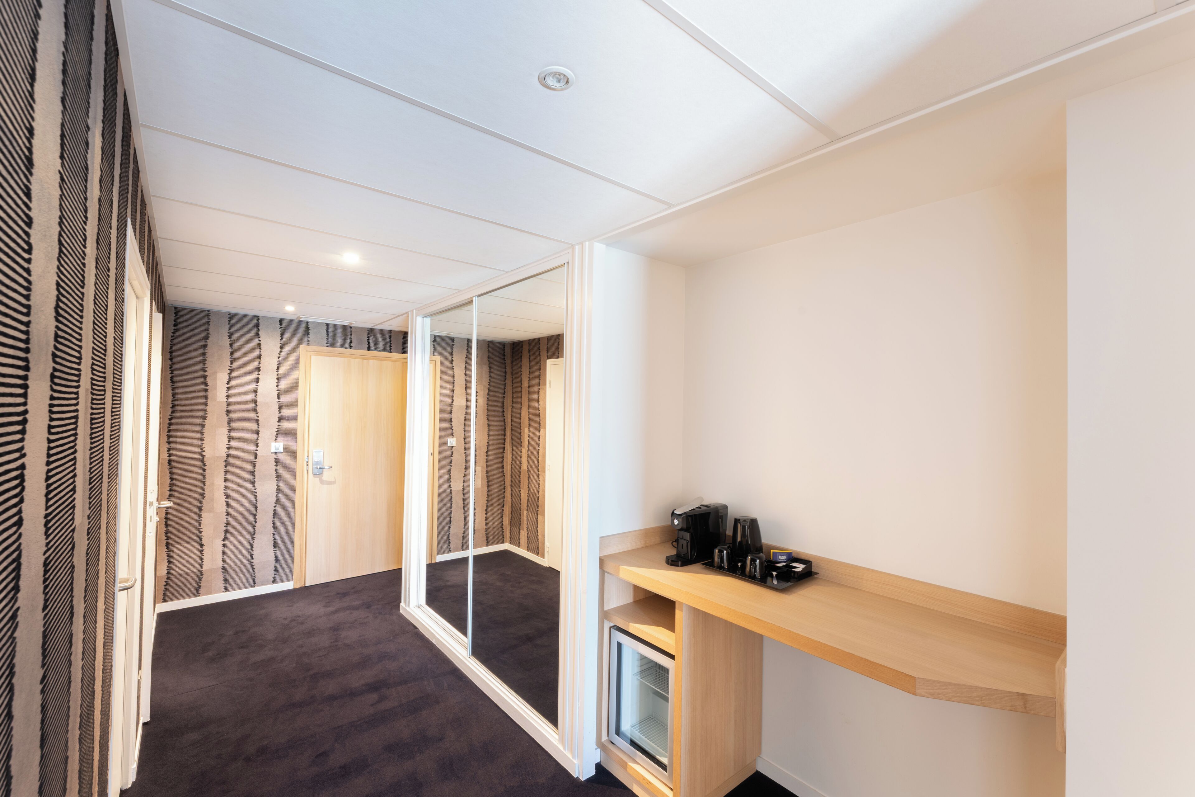 Quarto de hotel moderno com carpete escuro, uma escrivaninha de madeira com máquina de café e um espelho grande.