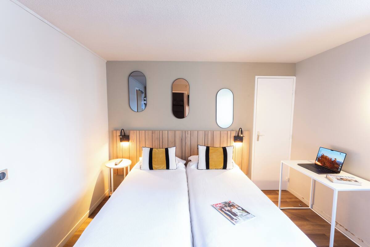 Quarto de hotel com duas camas, uma mesa com um laptop e três espelhos fixados na parede.