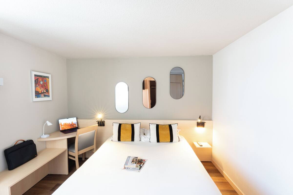 Quarto de hotel com cama, escrivaninha, cadeira, mesa de cabeceira, luminária e uma revista na cama.