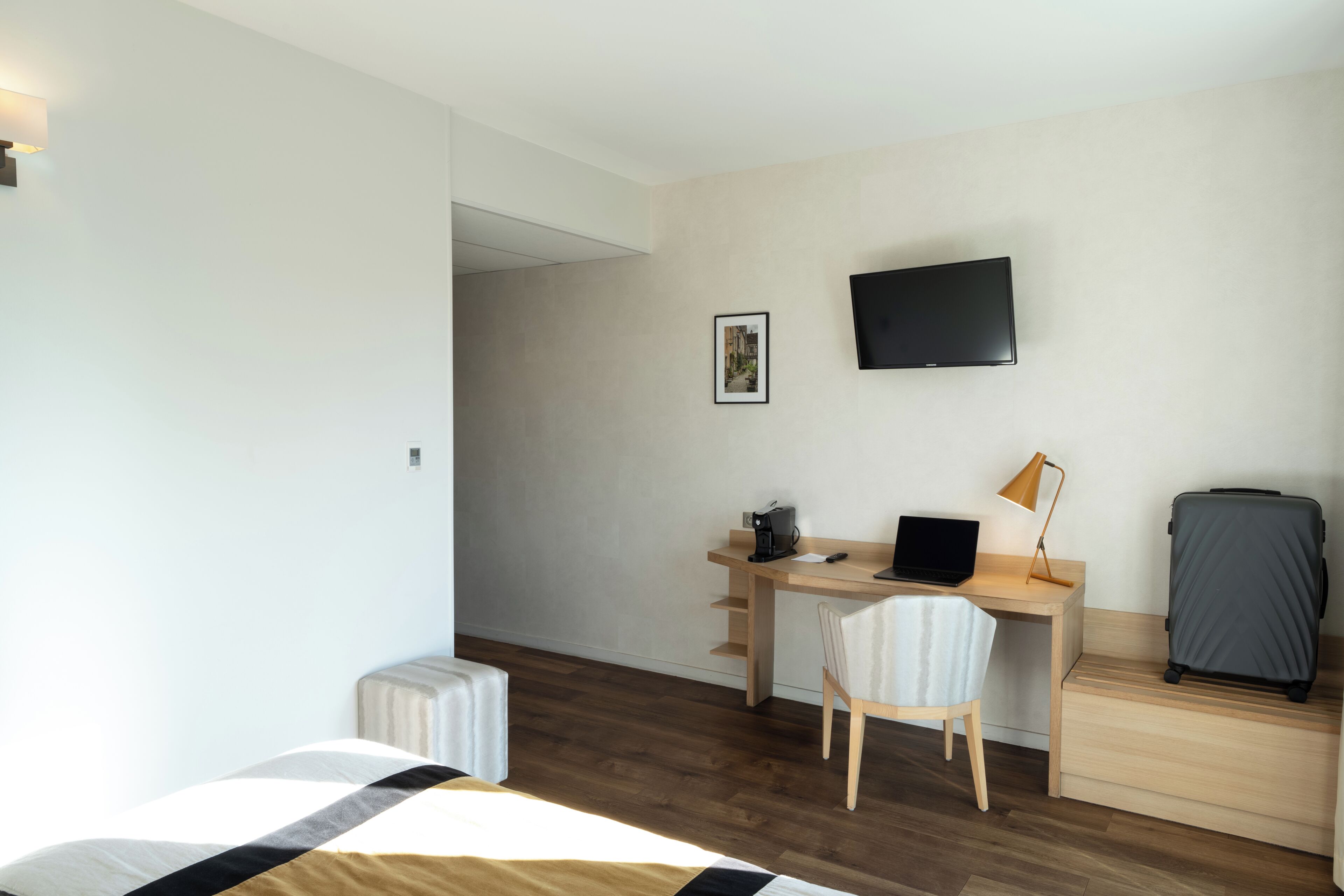 Una camera d'albergo con un letto, una scrivania con un portatile, una sedia, una televisione e una valigia.
