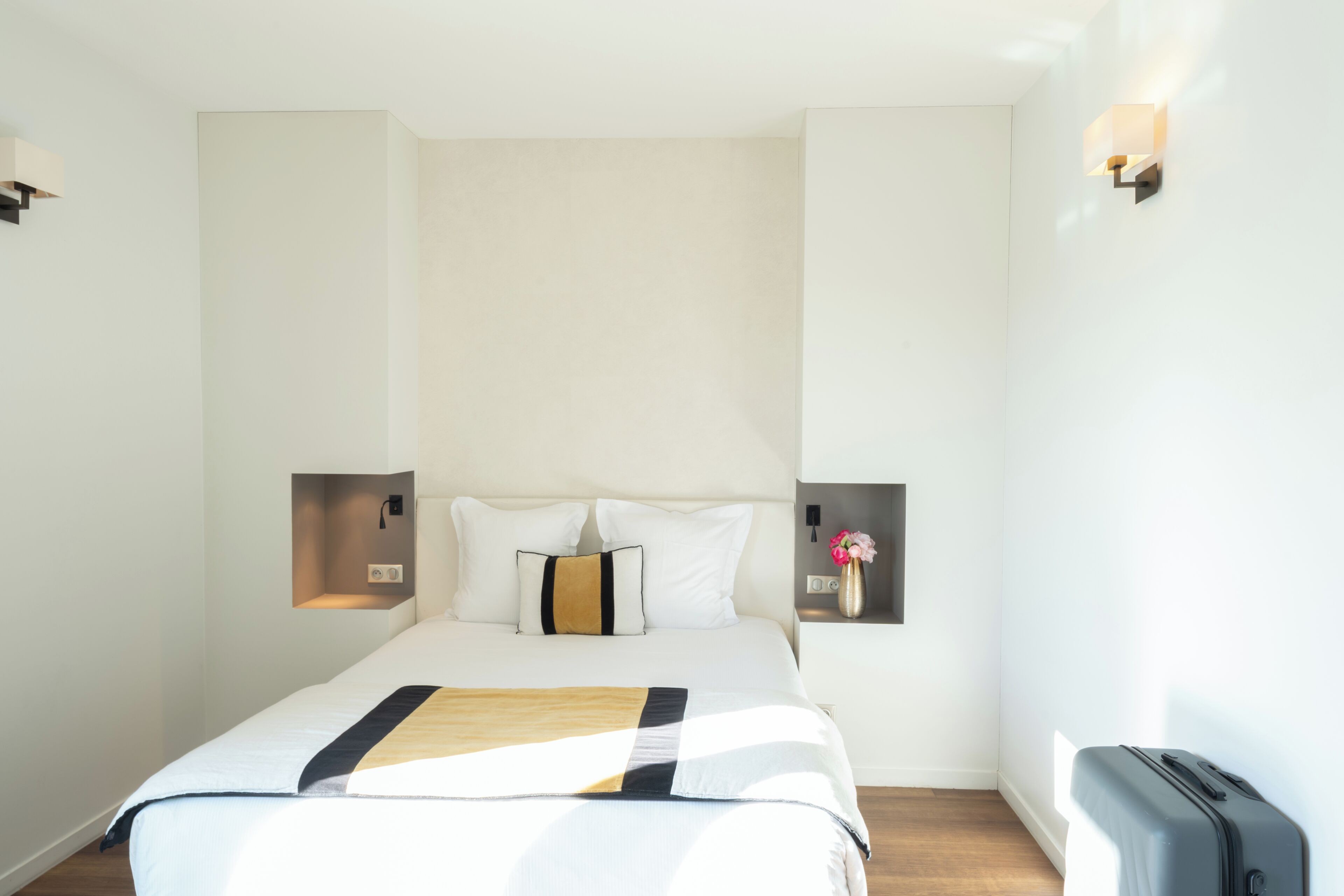Una camera d'albergo con un letto, comodini, una lampada a parete e una valigia.