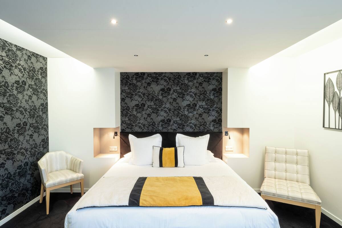 Quarto de hotel com uma cama grande, duas mesas de cabeceira, uma cadeira e cabeceira com padrão floral.