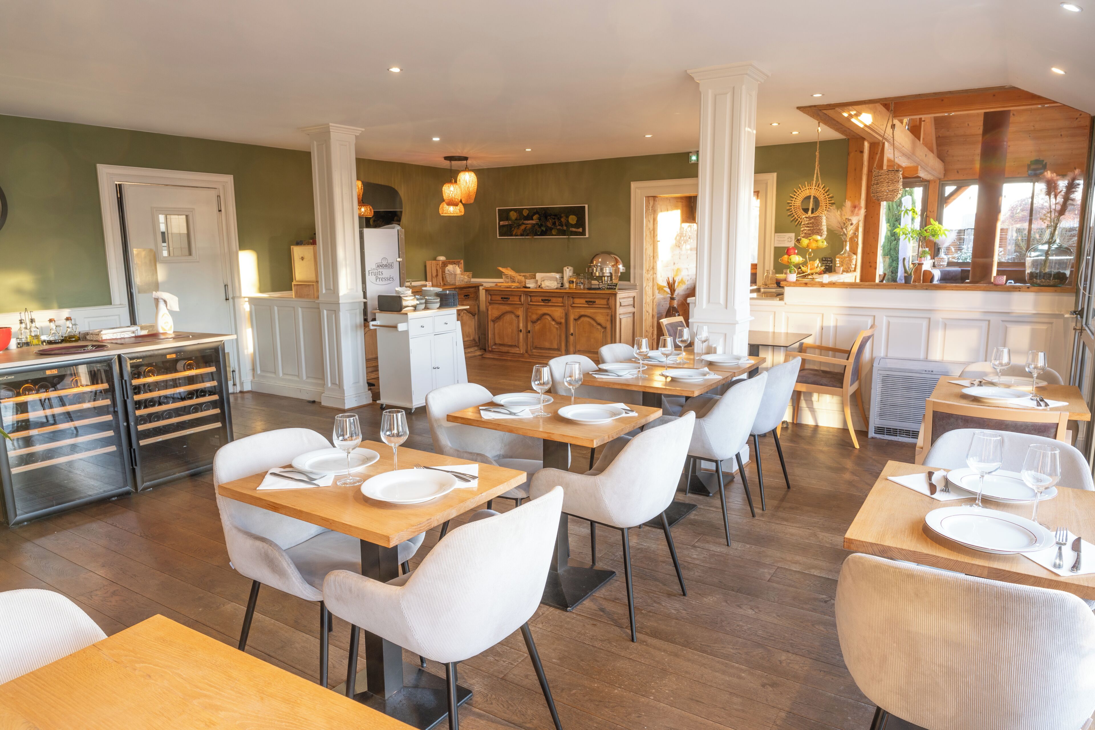 Comedor con mesas de madera y sillas blancas, una cocina al fondo y vistas al exterior a través de una ventana.