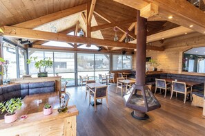 Un restaurante con vigas de madera, ventanas amplias y una estufa de leña en el centro.