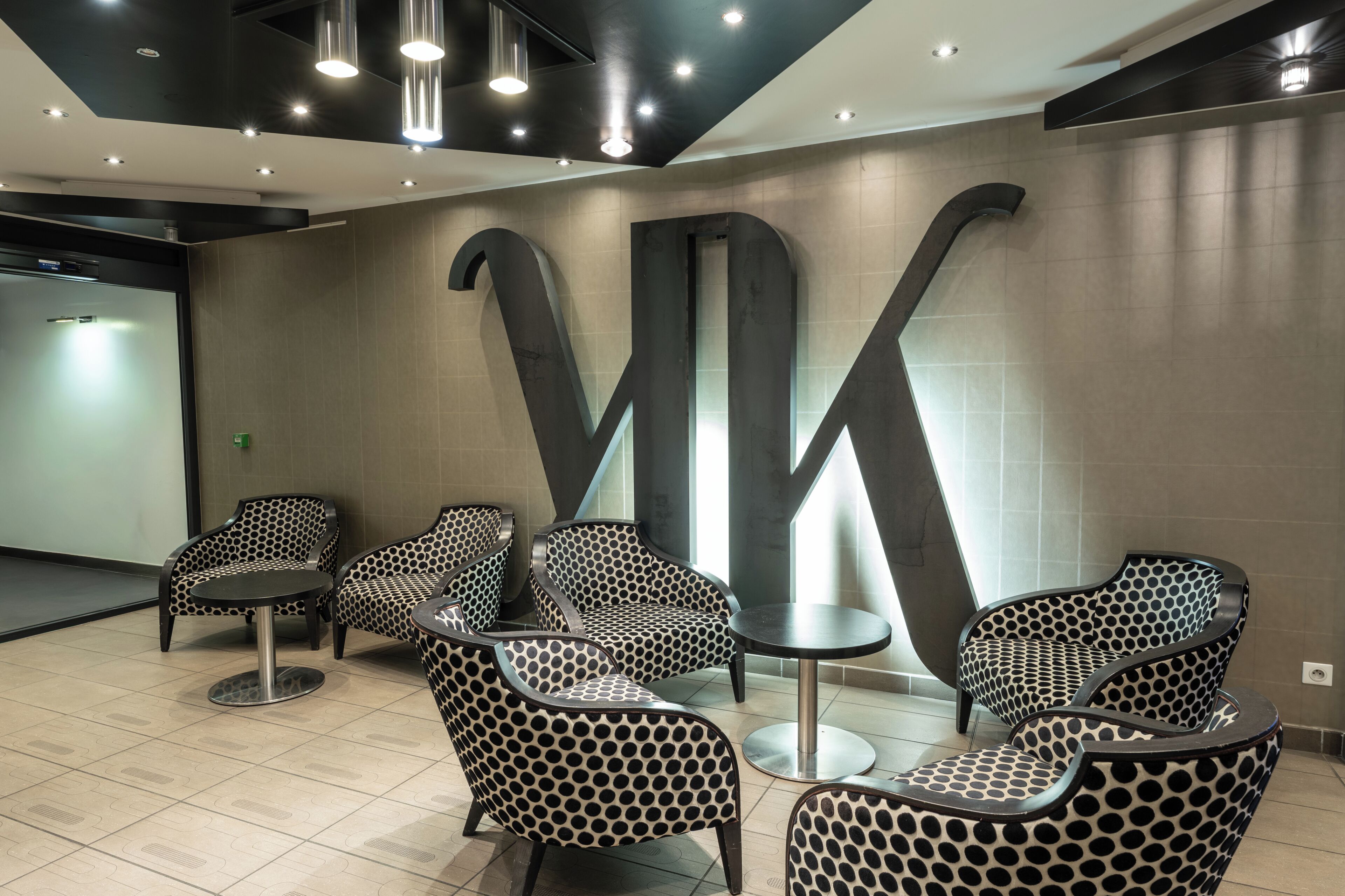 A modern waiting area with patterned chairs and tables, a large wall-mounted sign, and recessed lighting.