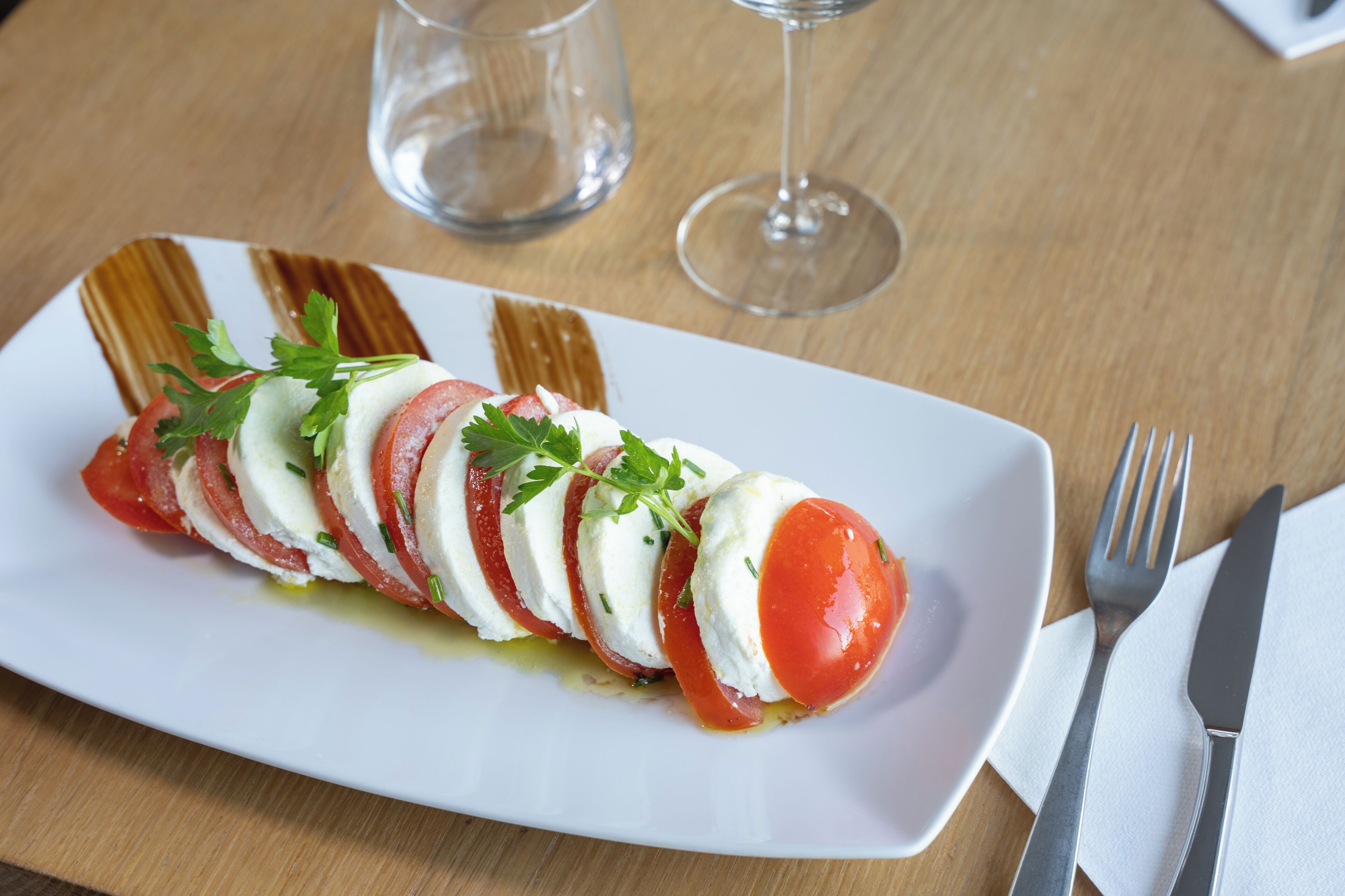 Un plato blanco con una ensalada de tomate y mozzarella, un tenedor y un cuchillo sobre una mesa de madera.